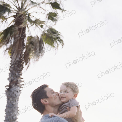 Man standing by a palm tree, carrying his young son in his arms, kissing him on the cheek.