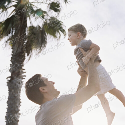 Man standing by a palm tree, playing with his young son, lifting him into the air.
