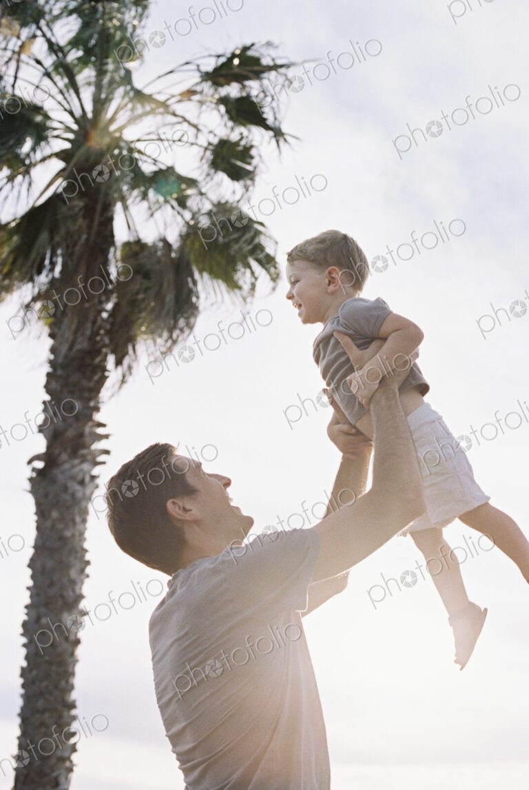 Man standing by a palm tree, playing with his young son, lifting him into the air.