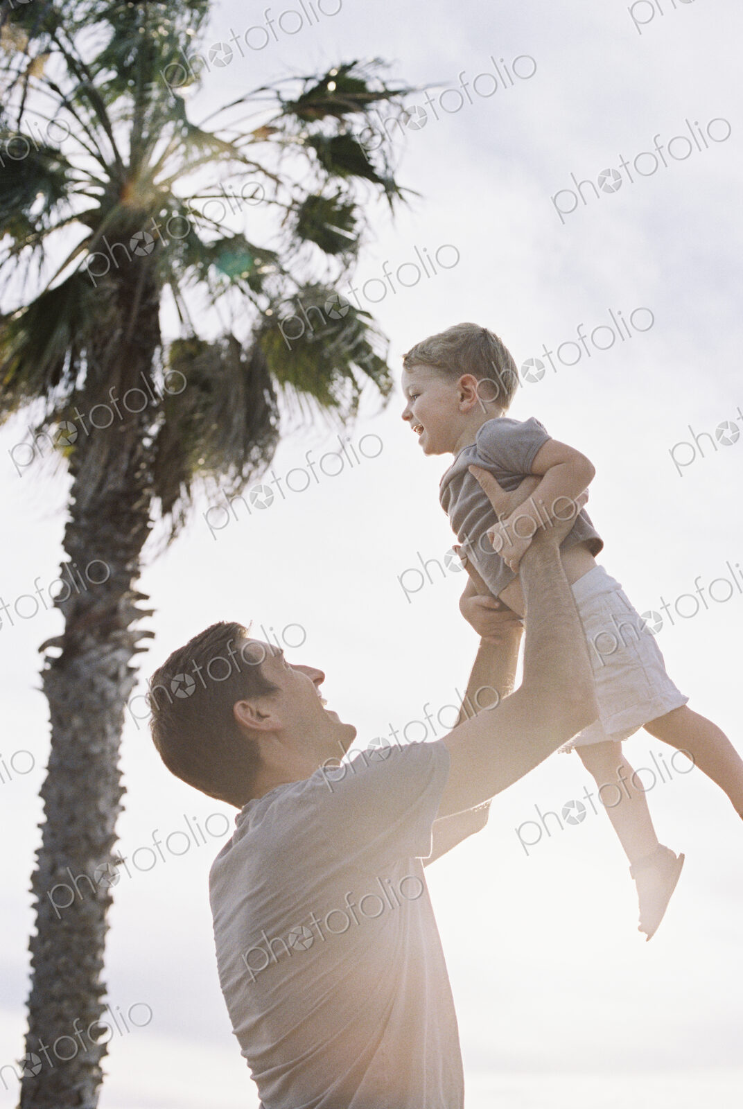Man standing by a palm tree, playing with his young son, lifting him into the air.