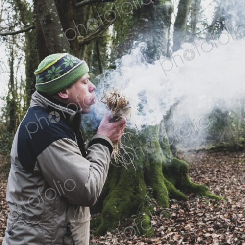 Man standing in a forest, blowing on bundle of straw, igniting fire.