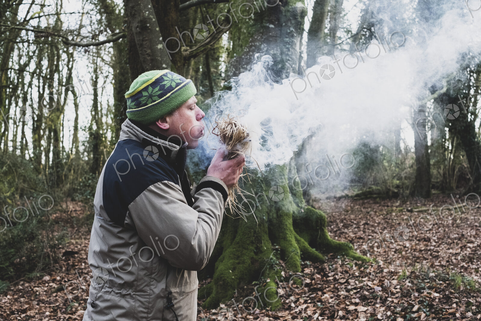 Man standing in a forest, blowing on bundle of straw, igniting fire.