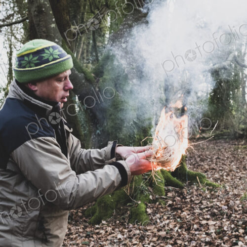 Man standing in a forest, holding lit-up bundle of straw, igniting fire.