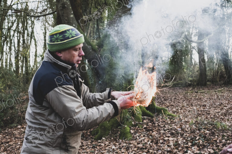 Man standing in a forest, holding lit-up bundle of straw, igniting fire.