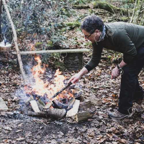 Man standing in a forest, stoking campfire, wooden tripod and cast iron metal pot.