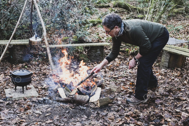 Man standing in a forest, stoking campfire, wooden tripod and cast iron metal pot.