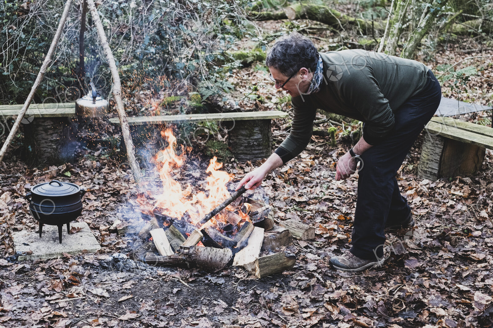 Man standing in a forest, stoking campfire, wooden tripod and cast iron metal pot.