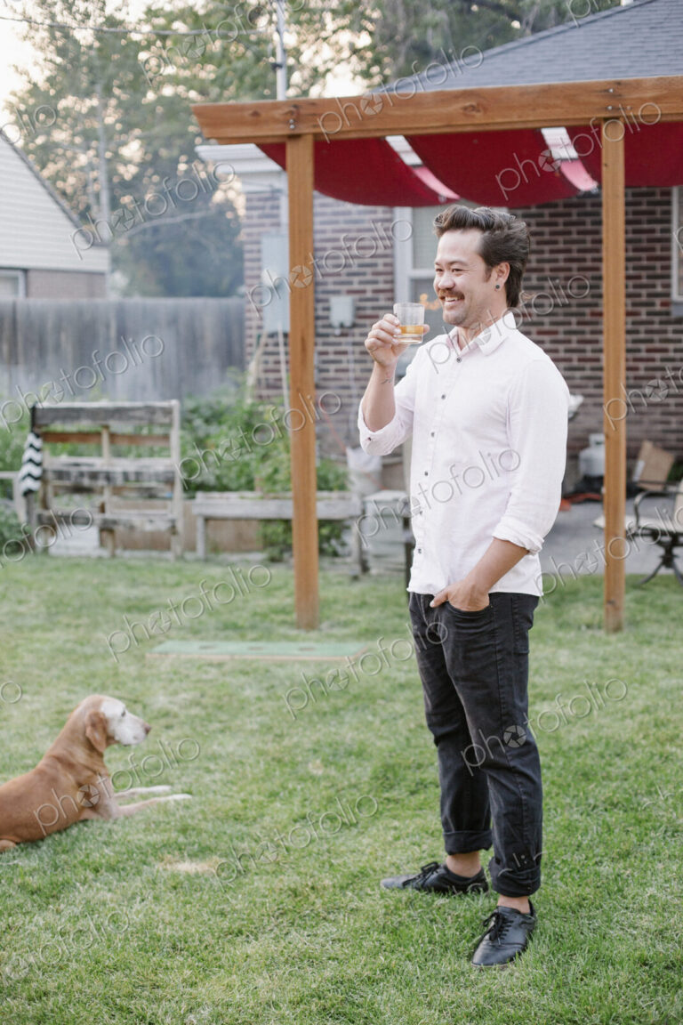 Man standing in a garden, enjoying a drink, a dog lying on the grass beside him.
