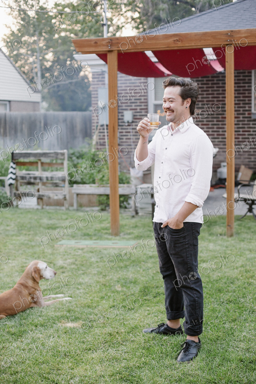 Man standing in a garden, enjoying a drink, a dog lying on the grass beside him.
