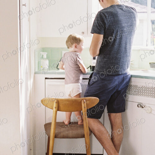 Man standing in a kitchen, his son standing on a chair beside him. Man standing in a kitchen, his son standing on a chair beside him.