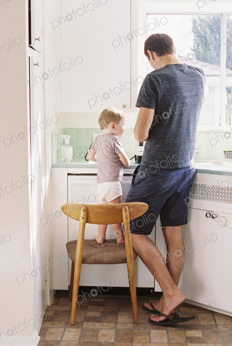 Man standing in a kitchen, his son standing on a chair beside him.