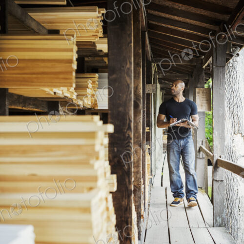 Man standing in a lumber yard, holding a folder, writing notes.