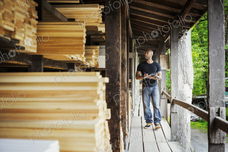 Man standing in a lumber yard, holding a folder, writing notes.