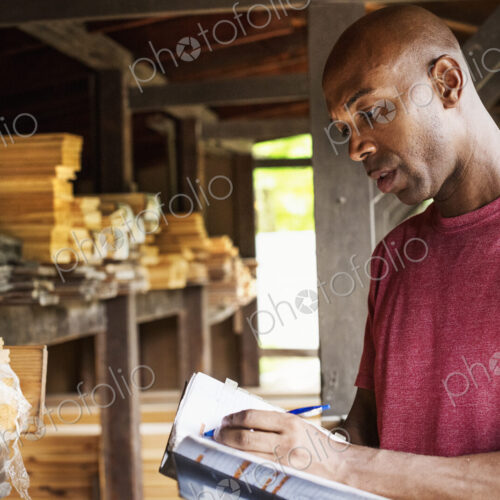Man standing in a lumber yard, holding a folder, writing notes.