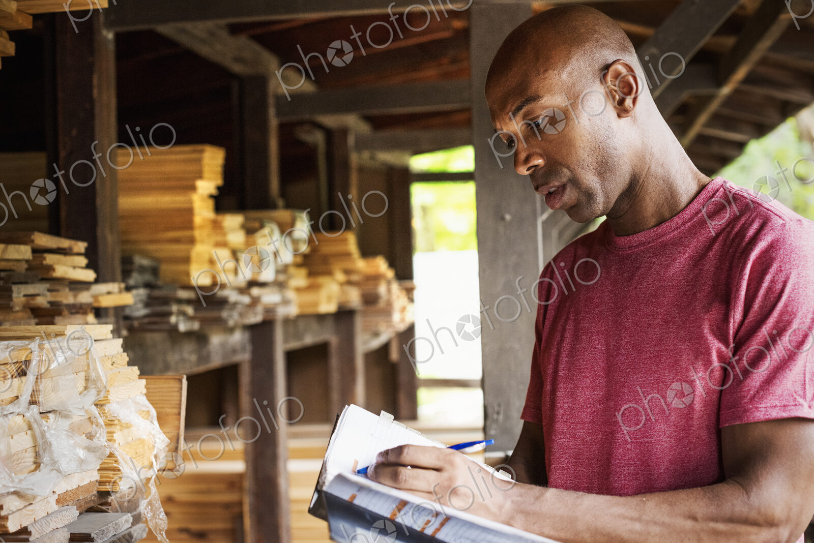 Man standing in a lumber yard, holding a folder, writing notes.