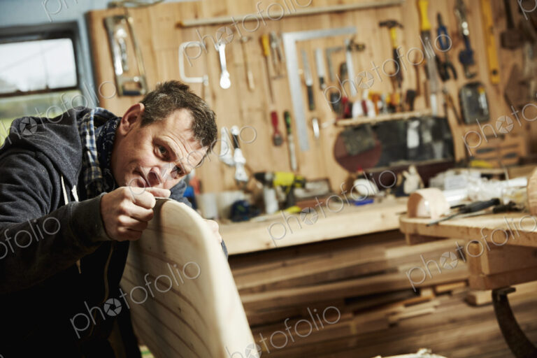 Man standing in a workshop inspecting closely the edge of a wooden surfboard.