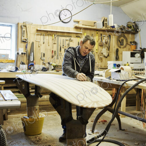 Man standing in a workshop sanding and shaping a wooden surfboard. Man standing in a workshop sanding and shaping a wooden surfboard.