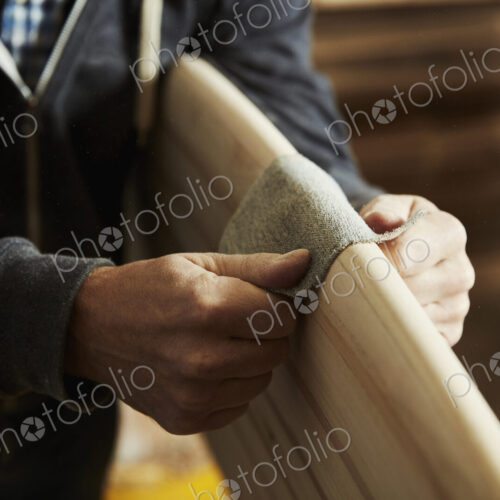 Man standing in a workshop sanding the rounded edge of a wooden surfboard. Man standing in a workshop sanding the rounded edge of a wooden surfboard.