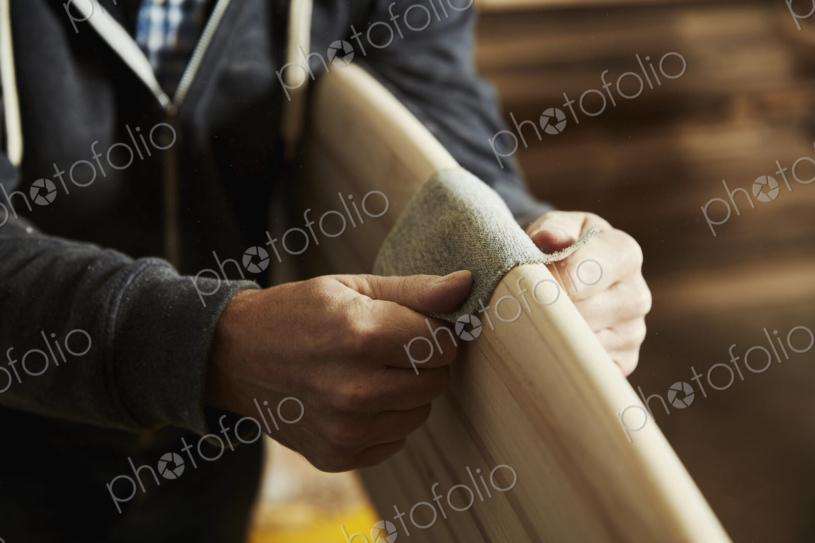 Man standing in a workshop sanding the rounded edge of a wooden surfboard.