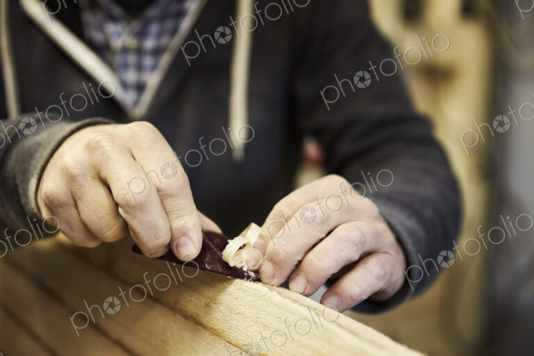 Man standing in a workshop, shaping a wooden surfboard using a hand held plane.