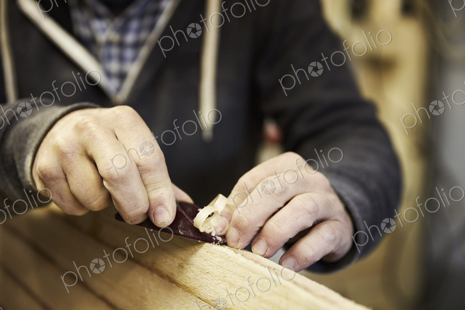 Man standing in a workshop, shaping a wooden surfboard using a hand held plane.