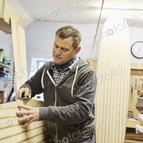 Man standing in a workshop, smoothing and shaping the surface of a wooden surfboard… Man standing in a workshop, smoothing and shaping the surface of a wooden surfboard…