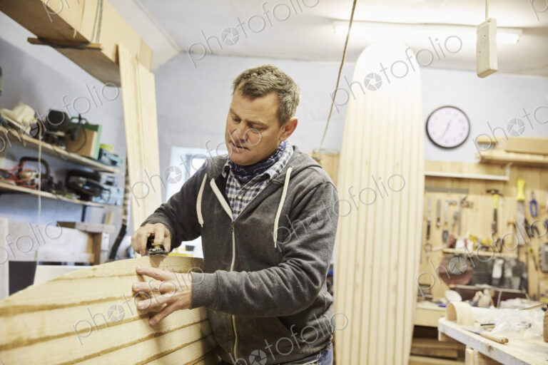Man standing in a workshop, smoothing and shaping the surface of a wooden surfboard…