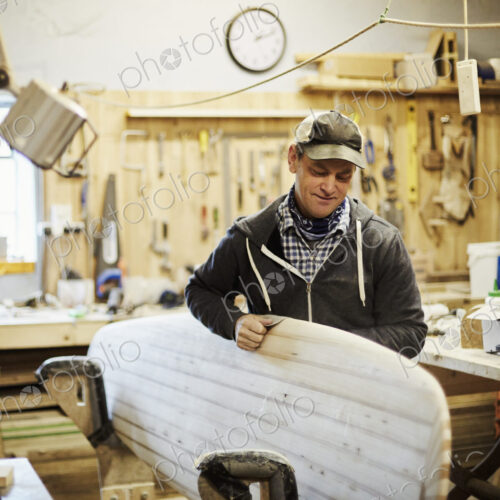 Man standing in a workshop smoothng and sanding a wooden surfboard. Man standing in a workshop smoothng and sanding a wooden surfboard.
