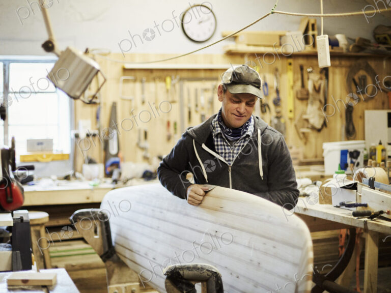 Man standing in a workshop smoothng and sanding a wooden surfboard.