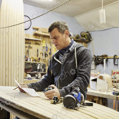 Man standing in a workshop with a wooden surf board, holding paper and pen. Man standing in a workshop with a wooden surf board, holding paper and pen.