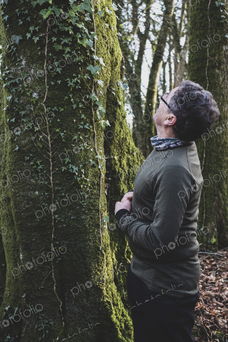 Man standing in forest, looking up tree overgrown with moss and ivy.