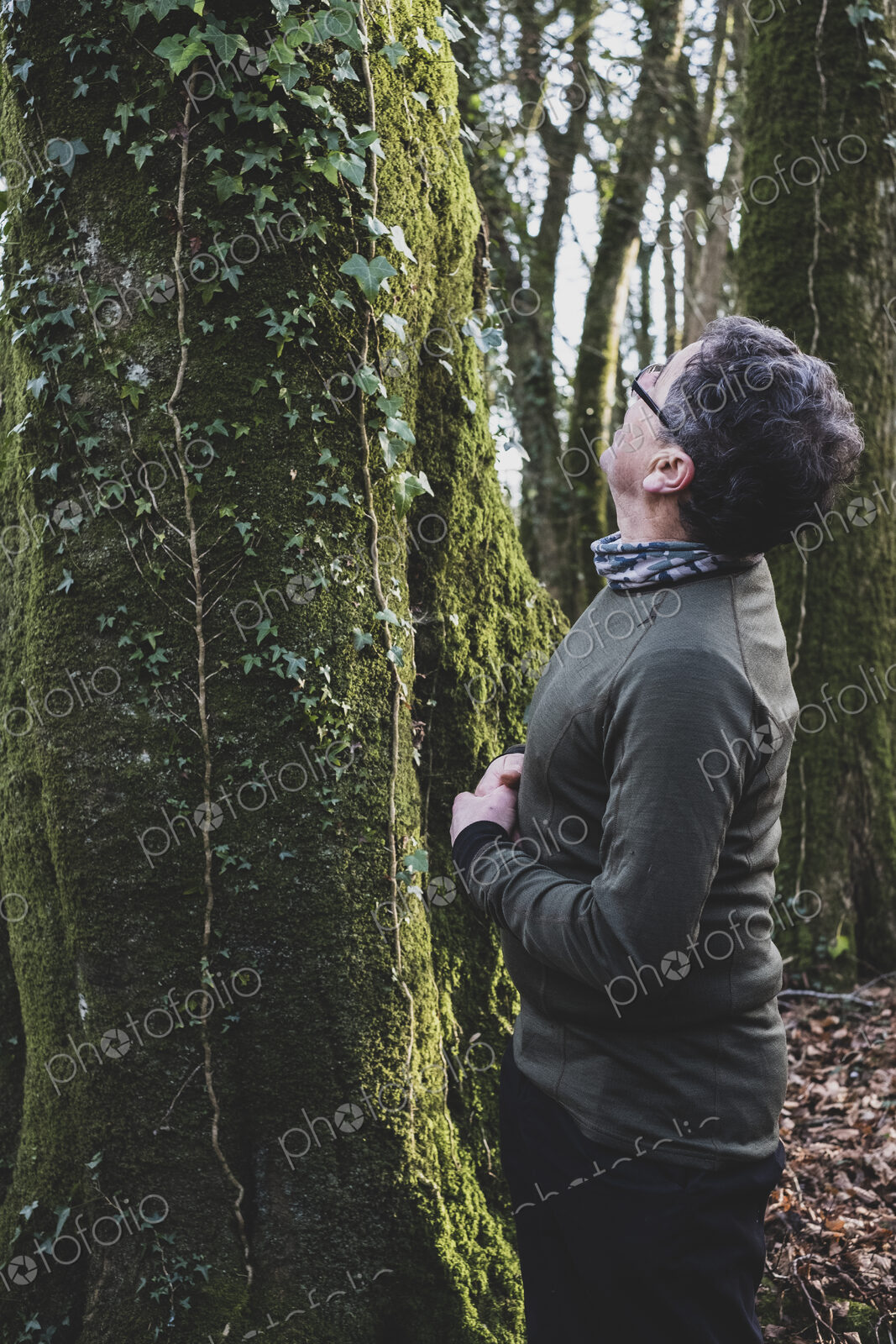 Man standing in forest, looking up tree overgrown with moss and ivy.