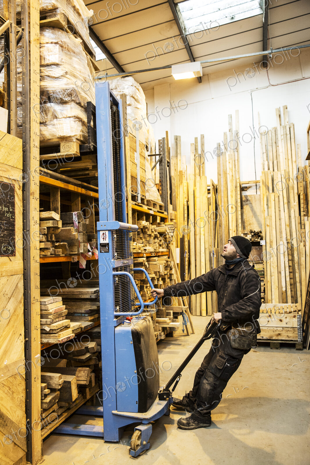 Man standing in warehouse using forklift to move pallet of wood off a shelf.