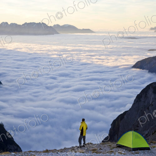 Man standing next to tent pitched above the clouds on a mountain summit.