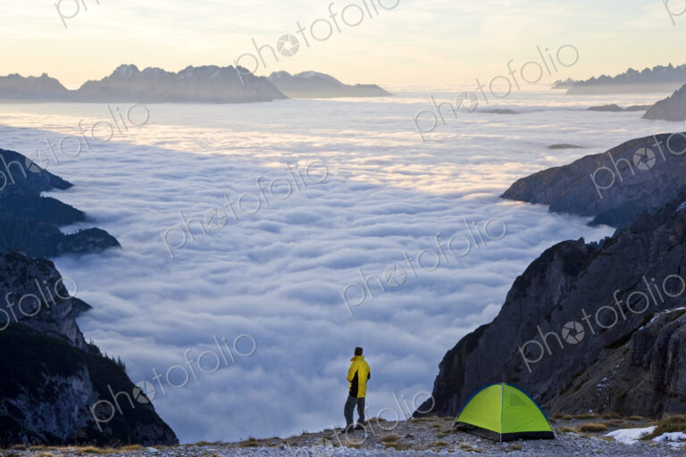 Man standing next to tent pitched above the clouds on a mountain summit.