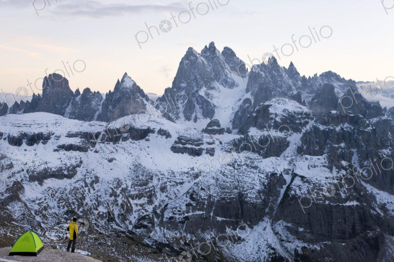 Man standing next to tent pitched amidst snow-covered mountain range.
