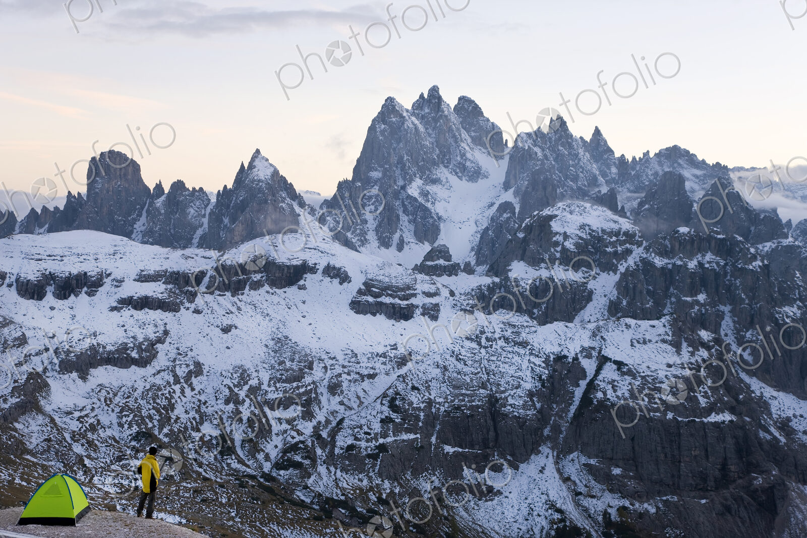 Man standing next to tent pitched amidst snow-covered mountain range.