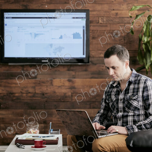 Man using a laptop balanced on his knee, sitting on sofa in a small office.