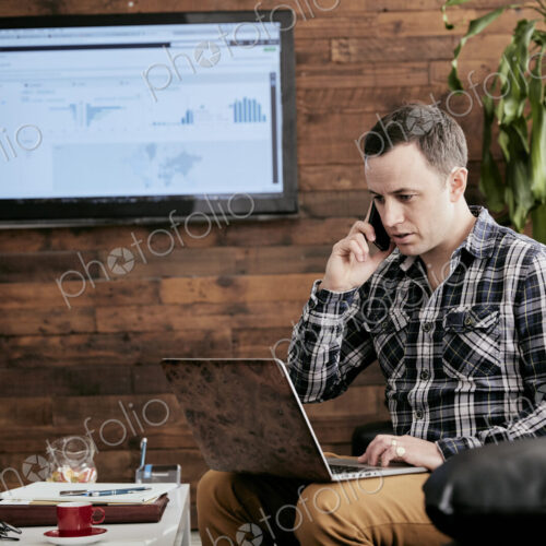 Man using a mobile phone using laptop sitting on sofa in a small office. Man using a mobile phone using laptop sitting on sofa in a small office.
