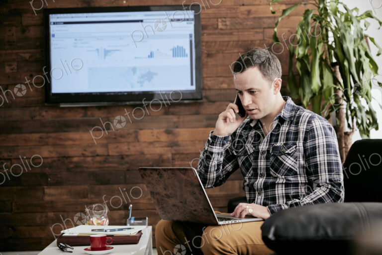Man using a mobile phone using laptop sitting on sofa in a small office.