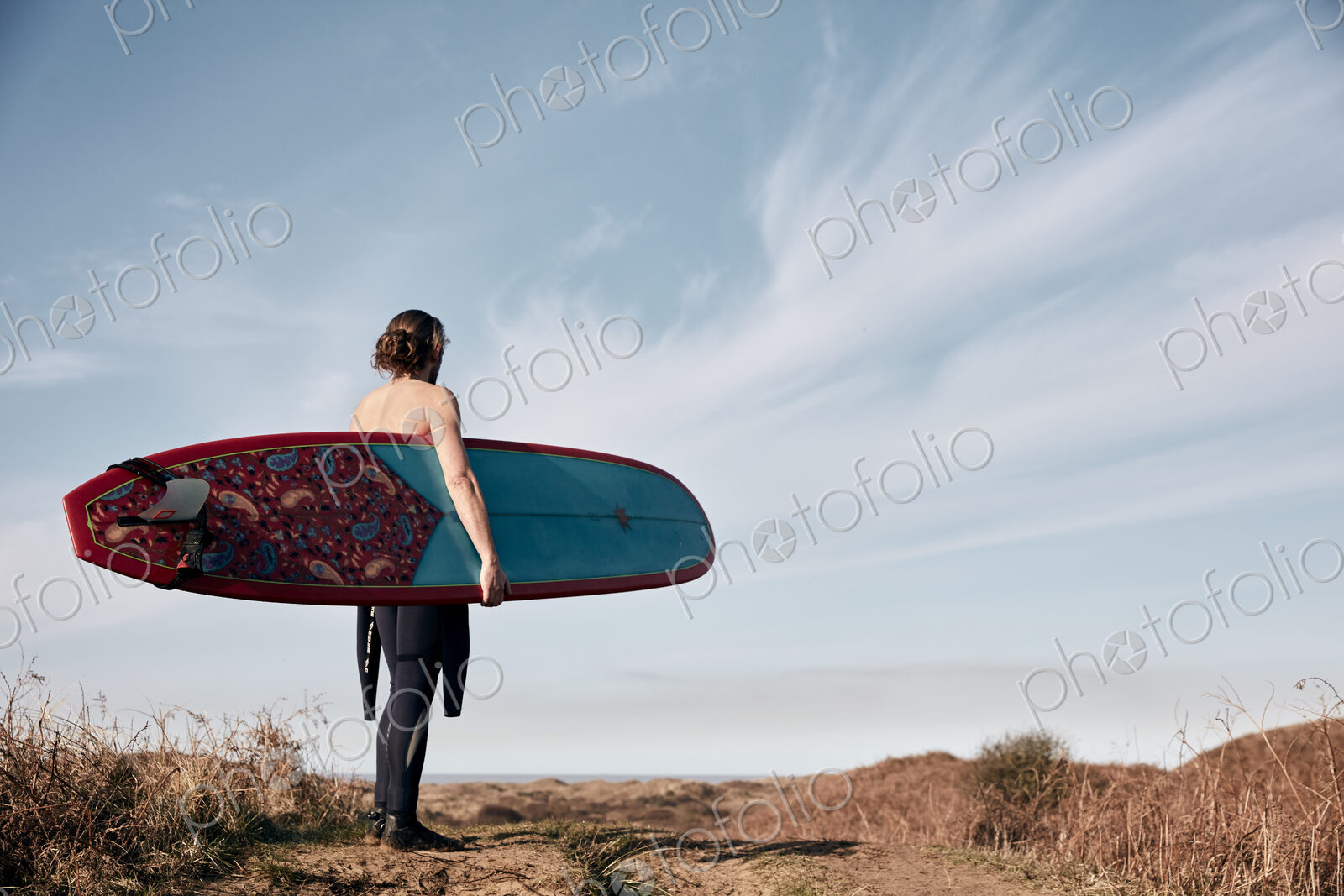 Man walking on a path in the sand dunes towards sea carrying a surf board.