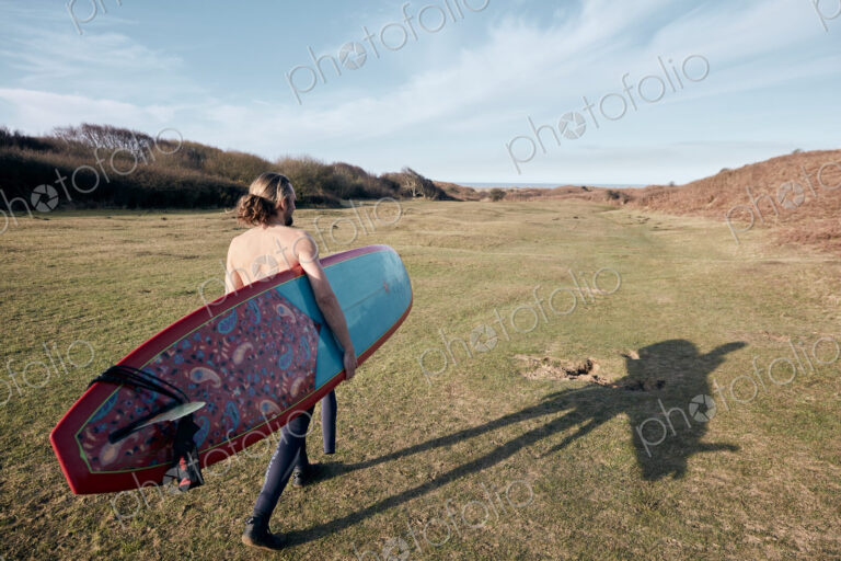 Man walking on a path in the sand dunes towards sea with a surf board