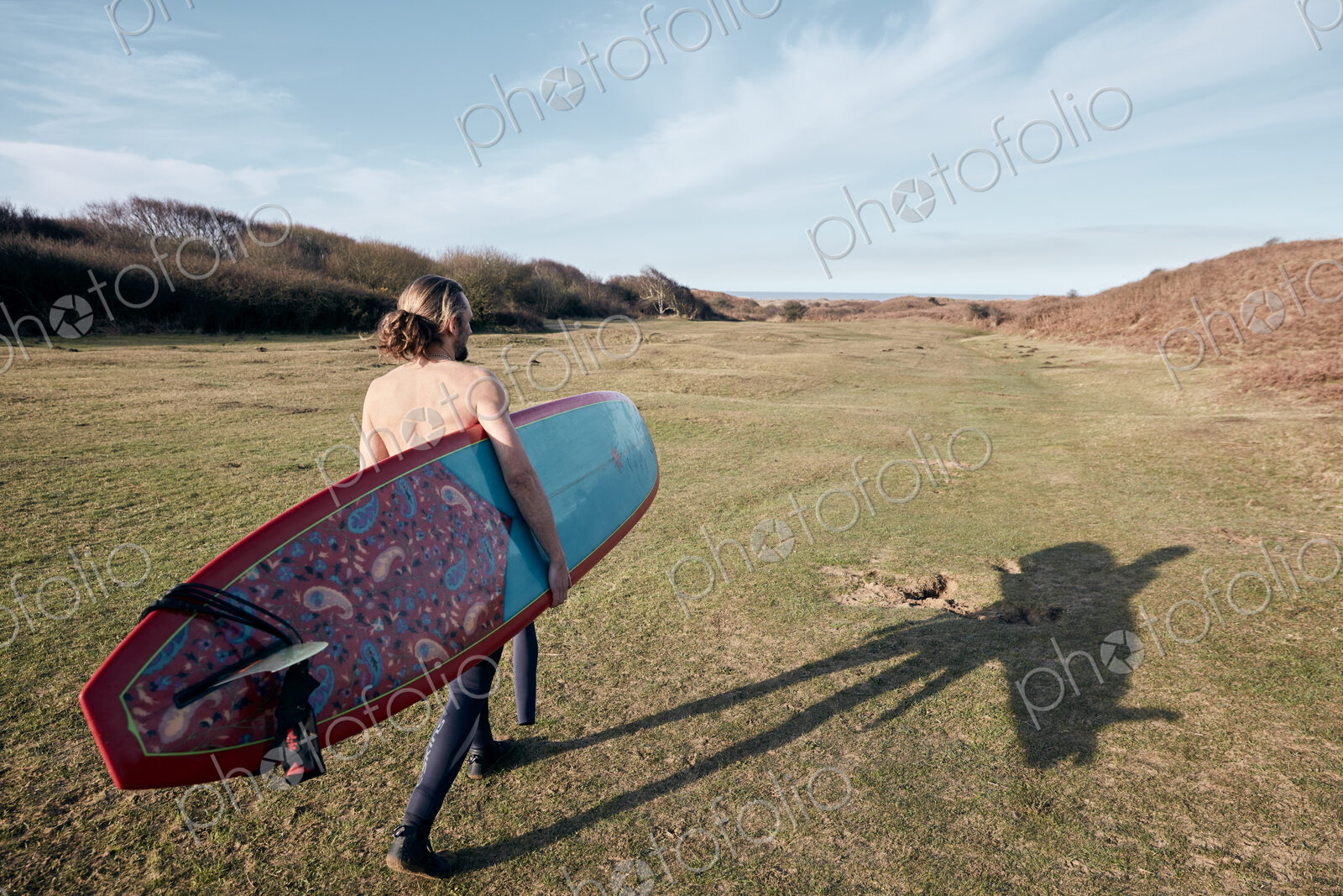 Man walking on a path in the sand dunes towards sea with a surf board