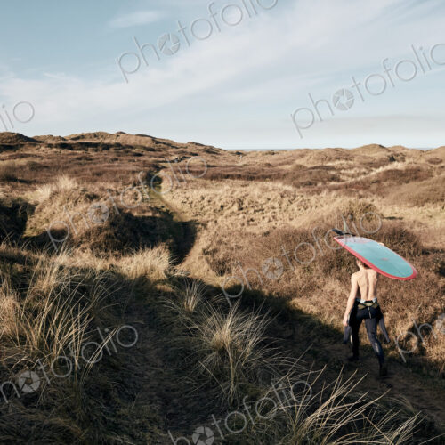 Man walking on a path in the sand dunes towards sea with surf board balancing on his head Man walking on a path in the sand dunes towards sea with surf board balancing on his head