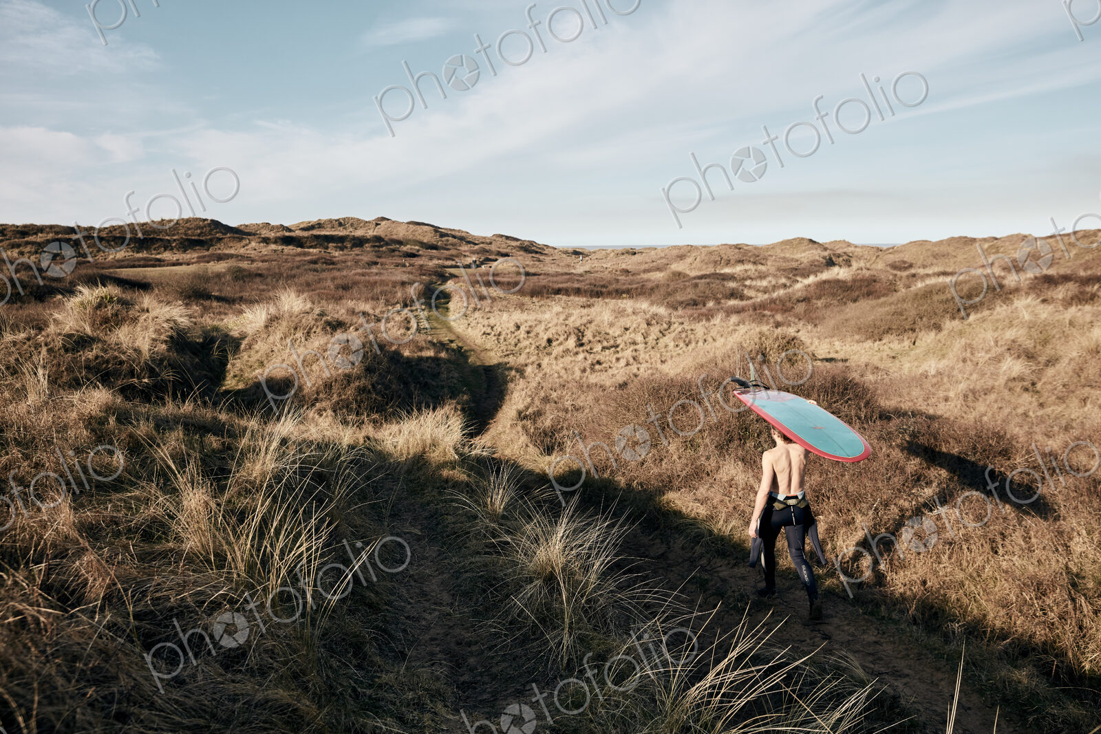 Man walking on a path in the sand dunes towards sea with surf board balancing on his head