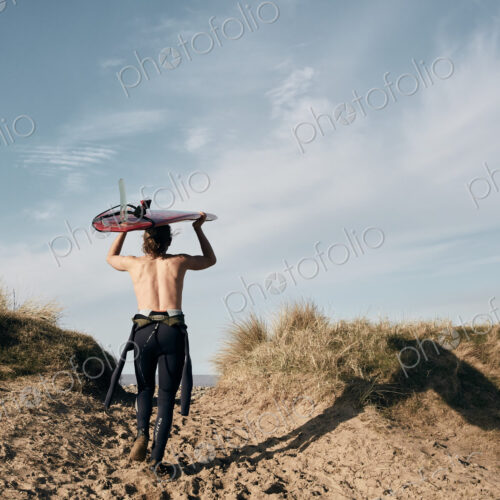 Man walking on a path in the sand dunes towards sea with surf board balancing on his head Man walking on a path in the sand dunes towards sea with surf board balancing on his head