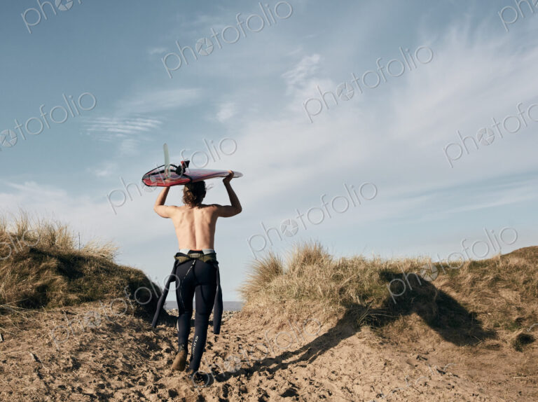 Man walking on a path in the sand dunes towards sea with surf board balancing on his head
