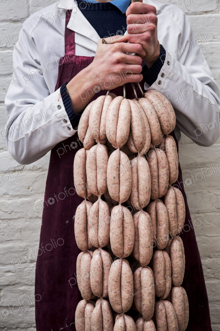 Man wearing apron holding large number of freshly made sausages hanging from hooks.