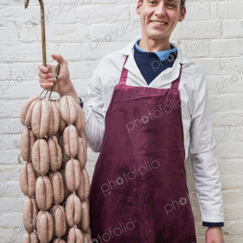 Man wearing apron holding large number of freshly made sausages hanging from hooks,… Man wearing apron holding large number of freshly made sausages hanging from hooks,…
