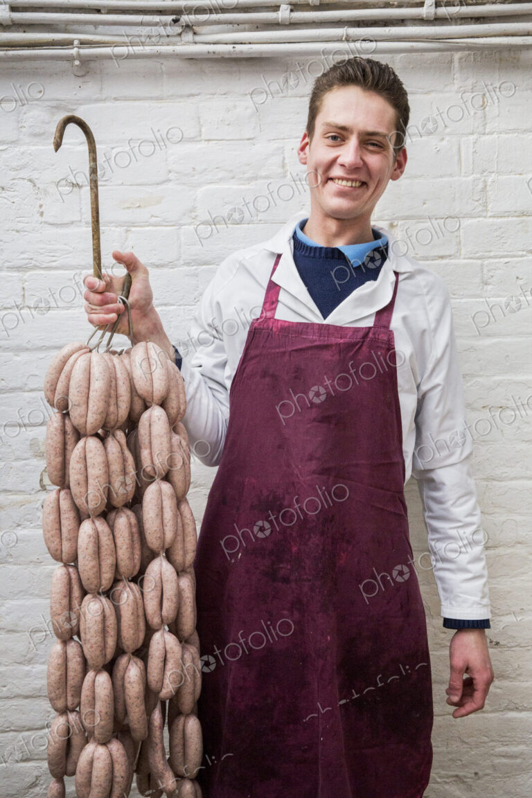 Man wearing apron holding large number of freshly made sausages hanging from hooks,…